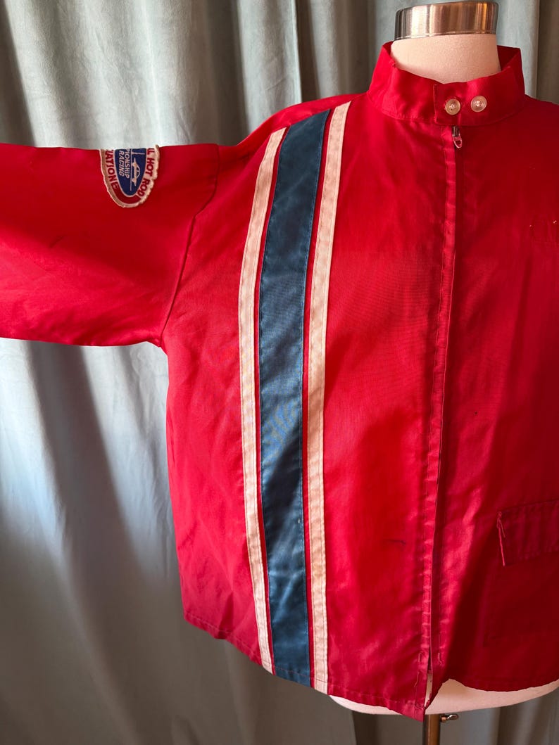 May include: A red racing jacket featuring a central blue stripe flanked by white stripes. The jacket has a high collar, a zip, and a patch on the sleeve with text. The jacket is a vintage style.