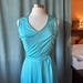 May include: A sleeveless turquoise dress featuring a V-neckline and a matching fabric tie belt at the waist. The garment is displayed on a mannequin and is constructed from a smooth, slightly reflective material with vertical seam detailing.