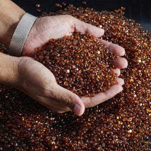 May include: A close-up shot of a pile of small, amber-colored glass beads. The beads are held in cupped hands, with a silver mesh bracelet visible on the wrist. The beads are smooth and reflective, creating a warm, inviting aesthetic.