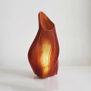 May include: A tall, sculptural table lamp with a deep orange, vertically ribbed exterior. The lamp features a fluid, asymmetrical silhouette that tapers toward the top, emitting a warm, glowing light from within its textured, pleated structure.