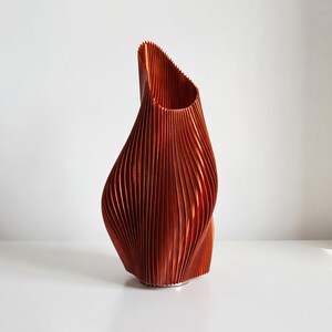 May include: A copper-colored, abstract vase with a textured, vertically-lined design. The vase has a unique, twisted shape and a wide opening at the top. The decorative piece is made of a ridged material.