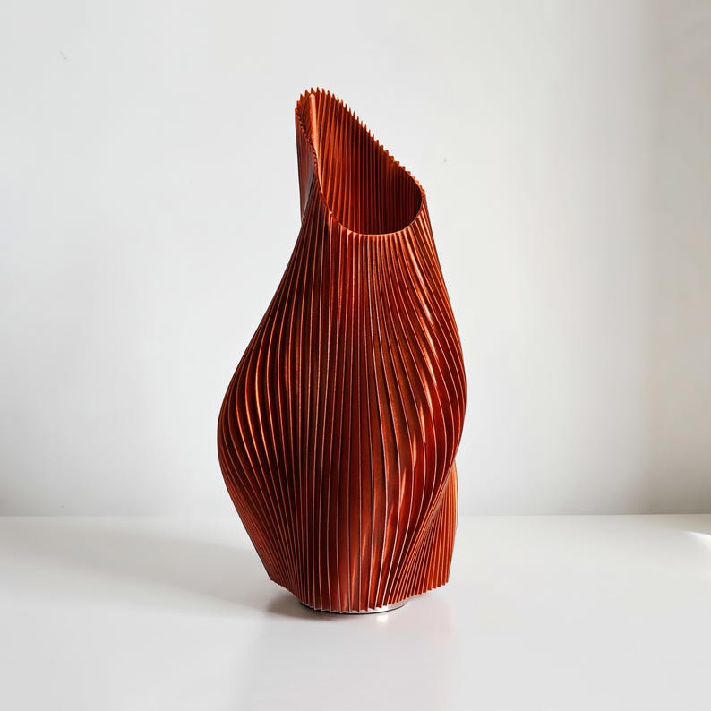 May include: A copper-colored, abstract vase with a textured, vertically-lined design. The vase has a unique, twisted shape and a wide opening at the top. The decorative piece is made of a ridged material.