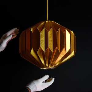 Puede incluir: Una lámpara colgante de estilo origami geométrico de color dorado cuelga del techo. La lámpara está hecha de un material de tela plisado.