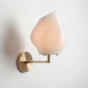 May include: A modern wall sconce with a white, textured shade and a brass-colored arm and base. The shade has a unique, organic shape with vertical lines. The sconce is mounted on a white wall, providing a clean and contemporary aesthetic.