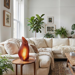 May include: A modern living room with a cream-colored sectional sofa, wooden side table, and a decorative amber-colored lamp. The lamp has a unique, curved design. The room is decorated with plants and framed artwork, creating a cozy and inviting atmosphere.