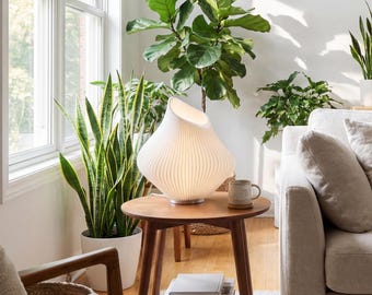 May include: A white, ribbed table lamp with a unique, asymmetrical shape sits on a small, round wooden side table. The lamp is turned on, illuminating the room. The table is surrounded by various potted plants and a sofa.