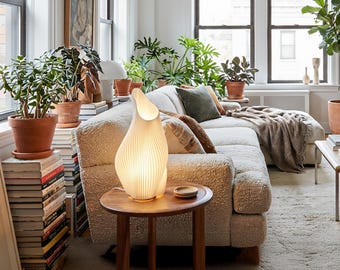May include: A modern living room scene featuring a cream-colored sofa, a wooden side table, and a unique, illuminated lamp. The lamp has a textured, organic design. Potted plants and books add to the cozy, inviting atmosphere.