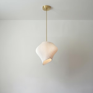 Op de afbeelding: Een moderne hanglamp met een witte, getextureerde kap en een messingkleurige plafondbevestiging. De kap heeft een unieke, gebogen vorm en een geribbeld ontwerp. Het licht hangt aan een dunne, messingkleurige staaf. De armatuur hangt tegen een witte achtergrond.
