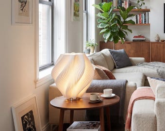 May include: A white, sculptural table lamp with a swirling, pleated design sits on a small, round, wooden side table. The lamp is illuminated, casting a warm glow. The table also holds two cups and saucers.