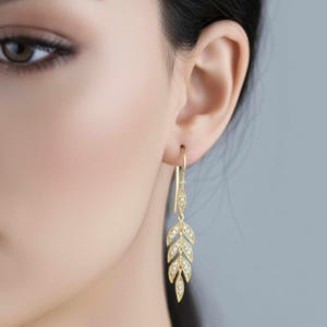 Puede incluir: Pendientes dorados con forma de hoja y pequeñas gemas transparentes. Los pendientes son colgantes y presentan un delicado diseño de hojas. Se lucen en una oreja, con un fondo suave y neutro.