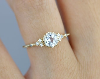 May include: A delicate gold ring with a round, clear gemstone surrounded by smaller, sparkling stones. The ring is set on a finger.