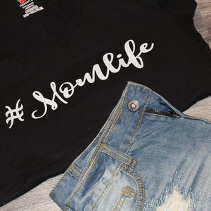 May include: A black t-shirt with a white hashtag and the text "#Momlife" in a cursive font. The t-shirt is paired with blue denim shorts.