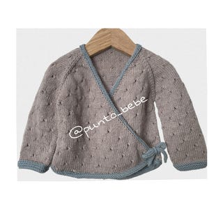 Puede incluir: Un cárdigan envolvente de punto gris con ribete azul y cierre de lazo. El cárdigan tiene un patrón texturizado y se muestra en una percha de madera. El texto "@punto_bebe" es visible.