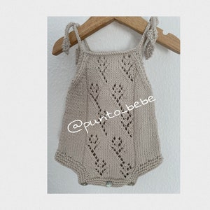 Puede incluir: Un body de bebé de punto beige con un delicado patrón de encaje y tirantes de lazo. El body tiene un nombre de perfil de redes sociales '@punto_bebe' impreso en la parte delantera.