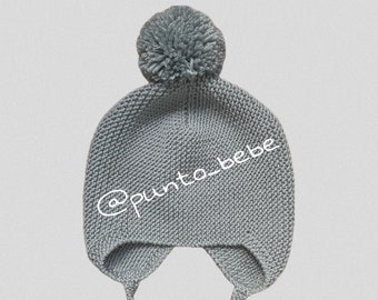 May include: Gray knitted baby hat with a pom pom on top and ear flaps tied with strings. The hat has the text "@punto_bebe" embroidered on the front.