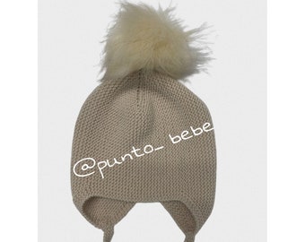May include: A beige knitted baby hat with a fluffy white pom pom and ear flaps tied with strings. The hat has the text "@punto_bebe" on the side.