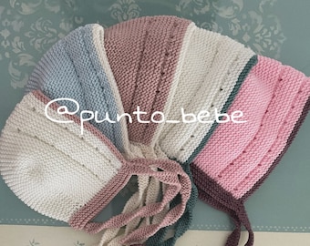May include: A knitted baby bonnet with a white, pink, light blue, and teal color scheme. The bonnet has a long tie that can be tied under the chin. The bonnet is made of soft yarn and is perfect for a baby girl.