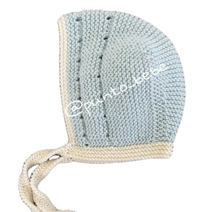 Puede incluir: Un gorro de bebé de punto azul claro con ribete blanco y una larga corbata blanca. El gorro tiene un patrón de pequeños agujeros.