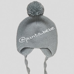 Puede incluir: Gorrito de bebé de punto gris con un pompón en la parte superior y orejeras atadas con cordones. El gorro tiene el texto "@punto_bebe" bordado en la parte delantera.