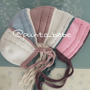 Puede incluir: Un gorro de punto para bebé con un esquema de color blanco, rosa, azul claro y turquesa. El gorro tiene una larga corbata que se puede atar debajo de la barbilla. El gorro está hecho de hilo suave y es perfecto para una niña.
