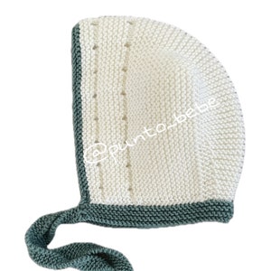 Könnte beinhalten: Eine weiße gestrickte Babyhaube mit grünem Besatz und einer langen grünen Schleife. Die Haube hat ein zartes Lochmuster.