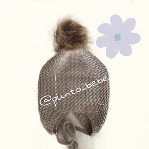 May include: A brown knitted baby hat with a fluffy brown pom pom on top. The hat has a tie at the bottom and the text '@punto_bebe' is embroidered on the side.