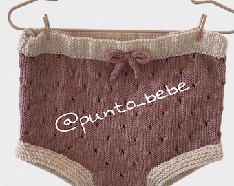 May include: A pair of knitted baby bloomers with a pink and cream color scheme. The bloomers have a bow at the waist and the text "@punto_bebe" is printed on the front.