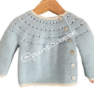 May include: A light blue knitted baby sweater with a white trim and button fastening. The sweater features a delicate stitch pattern and a slightly oversized fit.