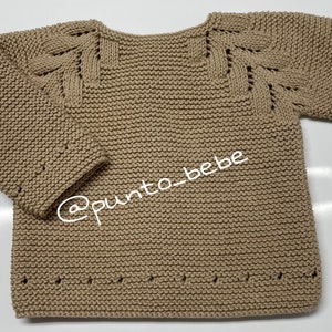 Puede incluir: Un suéter de punto beige con un patrón de punto único en los hombros y las mangas. El suéter tiene un cuello redondo y mangas largas. El suéter está hecho de un material suave y cómodo.