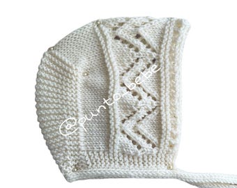 May include: Ivory knitted baby bonnet with a decorative zigzag pattern and small pearl accents. The bonnet has a tie closure and the text "@punto_bebe".
