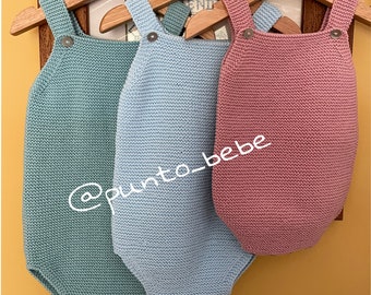 May include: Three knitted baby onesies hanging on wooden hangers. The onesies are in shades of light blue, teal, and pink. The onesies have straps and a button closure. The text '@punto_bebe' is visible on the image.