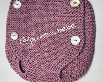 May include: A mauve knitted baby diaper cover with four white pearl buttons. The diaper cover is made with a textured knit stitch. The text '@punto_bebe' is visible on the diaper cover.