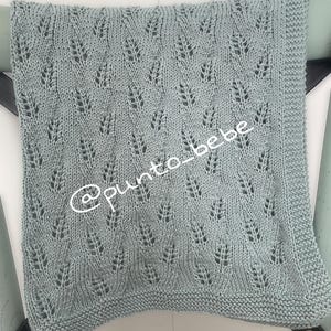 Puede incluir: Una manta de bebé de punto azul claro con un delicado patrón de hojas. La manta es rectangular y tiene una superficie texturizada. El texto "@punto_bebe" está escrito en la manta.