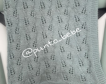 May include: A soft, light blue knitted baby blanket with a delicate leaf pattern. The blanket is rectangular and has a textured surface. The text "@punto_bebe" is written on the blanket.