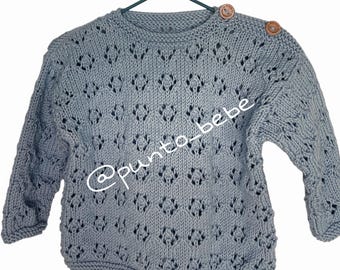 May include: A light blue long-sleeve knit sweater featuring an intricate openwork diamond pattern throughout the body and sleeves. The garment includes two round wooden buttons on the left shoulder and the text @punto_bebe across the front.