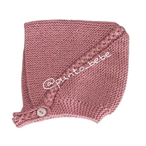 Puede incluir: Un gorro de bebé de punto rosa con un botón y un detalle de punto decorativo. @punto_bebe está escrito en el gorro.