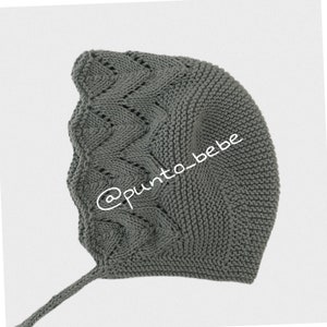 Könnte beinhalten: Eine graue gestrickte Babyhaube mit langem Band. Die Haube hat ein Zopfmuster und ein zartes Lochmuster. Der Text '@punto_bebe' ist auf der Haube sichtbar.