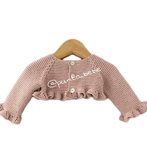 Puede incluir: Un cárdigan de bebé de punto rosa con un dobladillo y mangas con volantes. El cárdigan tiene un cierre de botón en la espalda.