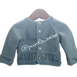 Puede incluir: Un cárdigan de punto azul claro para bebé con cierre de botones y un ribete de encaje decorativo. El cárdigan está colgado en una percha de madera. El texto "@punto_bebe" es visible en el cárdigan.