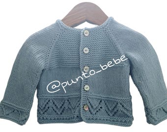 May include: A light blue knitted baby cardigan with a button closure and a decorative lace trim. The cardigan is hanging on a wooden hanger. The text "@punto_bebe" is visible on the cardigan.