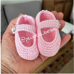 以下が含まれることがあります： 白いボタンのついたピンクのニットのベビーシューズのペア。靴はシンプルなデザインで、新生児に最適です。