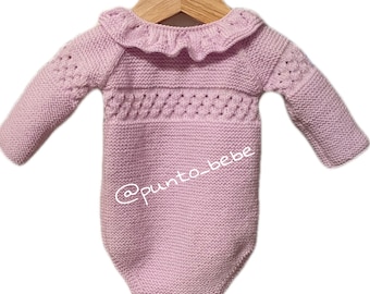 May include: A lavender knitted baby onesie with a ruffled collar and a delicate openwork pattern. The onesie is hanging on a wooden hanger. The text "@punto_bebe" is visible on the onesie.