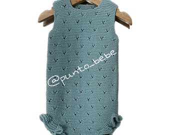 May include: A light blue knitted baby romper with a delicate openwork pattern and ruffled trim. The romper is hanging on a wooden hanger. The text '@punto_bebe' is visible on the romper.