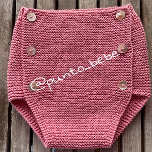 Puede incluir: Un pañal de bebé de punto rosa con cinco botones blancos. El pañal está hecho con un punto de tejido simple y tiene un diseño de pliegue.