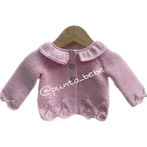 Op de afbeelding: Een roze gebreide babytrui met een witte kraag en knoopsluiting. De trui heeft een gegolfde zoom en hangt aan een houten hanger.