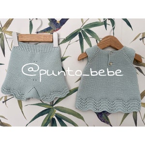 Puede incluir: Un conjunto de vestido y pantalones cortos de punto azul claro para bebé con ribete festoneado. El conjunto está colgado en perchas de madera sobre un fondo de papel tapiz estampado en verde y blanco.