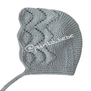 Może przedstawiać: Szary, dzianinowy czepek dla niemowlęcia z długim troczkiem. Czepek ma delikatny, ażurowy wzór i jest wykonany z miękkiej przędzy.