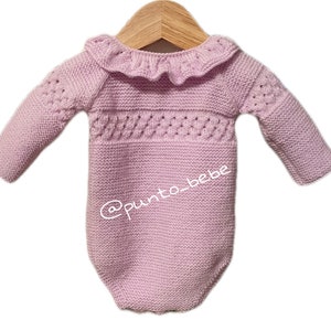 Könnte beinhalten: Ein helllila gestrickter Baby-Strampler mit langen Ärmeln und einem Rüschenkragen. Der Strampler hat ein dekoratives Strickmuster auf der Brust und wird auf einem Holzbügel präsentiert. Der Text "@punto_bebe" ist sichtbar.