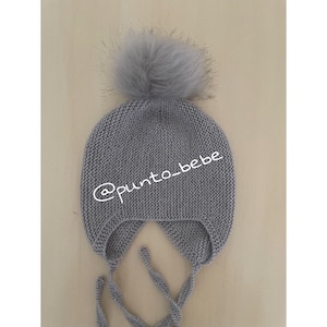 Könnte beinhalten: Graue gestrickte Baby-Mütze mit einem flauschigen Bommel oben und Bändern, die unter das Kinn gehen. Die Mütze hat den Text "@punto_bebe" an der Seite.