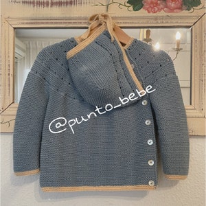 May include: A light blue knitted baby cardigan with a matching hood, featuring a button closure and a beige trim. The cardigan is hanging on a wooden hanger.